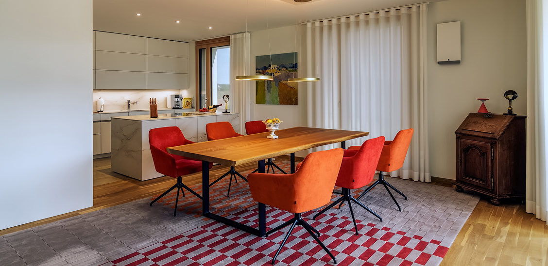 Moderne Esszimmer mit einem großen Holztisch, orangefarbenen Stühlen und offener Küche in einem Privathaus in Süddeutschland.
