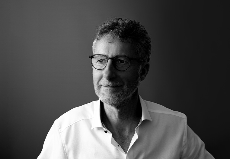 Ein lächelnder Mann mit Brille und grauen Haaren, der in einem hellen Hemd vor einem dunklen Hintergrund sitzt.